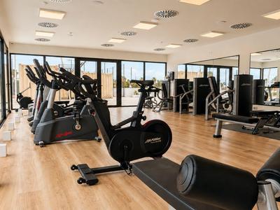 Helles Fitnessstudio mit Holzboden, Trainingsgeräten und großen Fenstern mit Ausblick.
