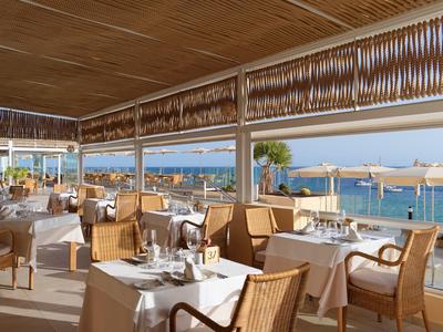 Offenes Restaurant mit Holzdecke, weißen Tischtüchern und Blick auf das blaue Meer und Palmen.