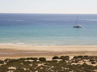 Calm sea with a sailboat near a sandy beach and sparse vegetation in the foreground.