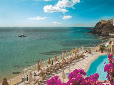 Beach resort with pool, loungers, and view of sea and cliffs under partly cloudy sky.