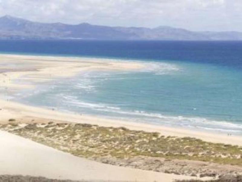 Wide sandy beach with calm blue sea, sparse vegetation, and distant mountains under a clear sky.