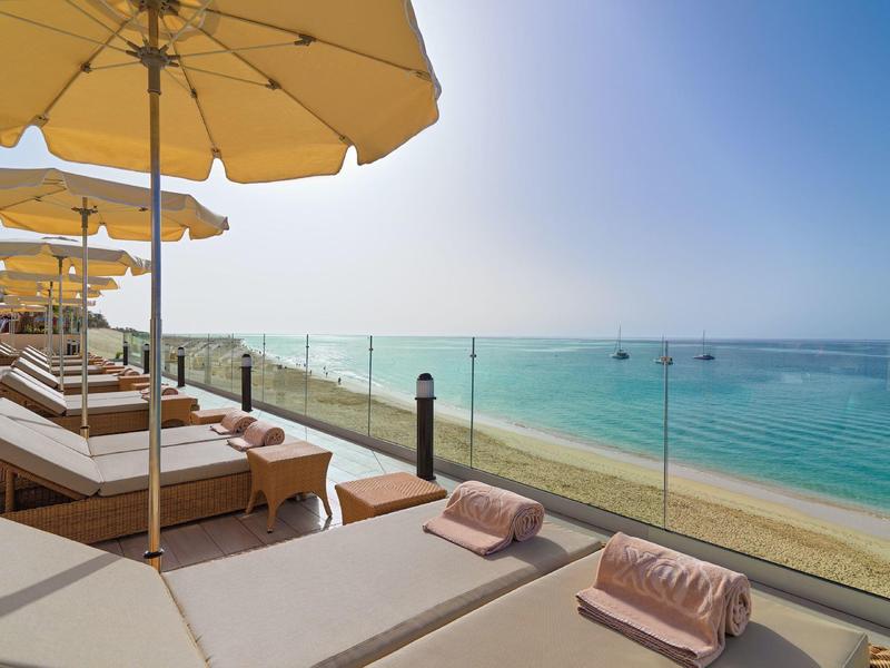 Beachfront lounge chairs under umbrellas on a terrace overlooking clear blue sea and sandy beach.