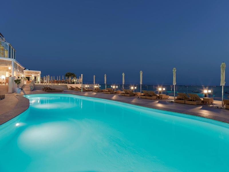 Illuminated outdoor pool at night with lounge chairs and ocean view at a seaside hotel.