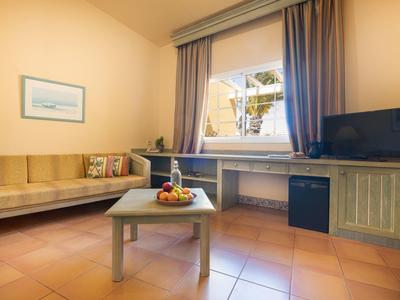 Camera d'hotel luminosa con divano ad angolo, tavolo con cesto di frutta e TV accanto a finestra con tende.