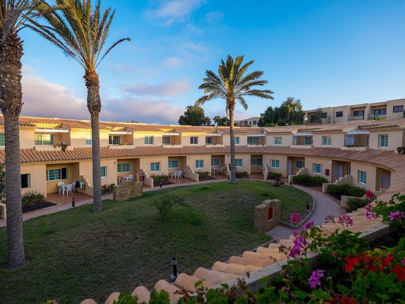 Complesso alberghiero con edifici a due piani, giardino di palme e piante in fiore al tramonto.