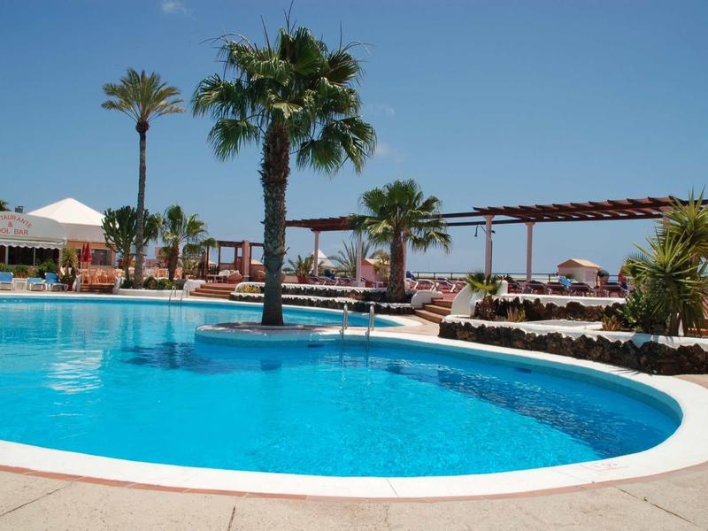 Runder Pool mit klarem Wasser, Palmen und Liegestühlen vor blauem Himmel am Strand.