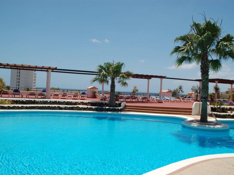 Großer blauer Pool mit Palmen und Sonnenliegen unter klarem Himmel an einem sonnigen Tag.