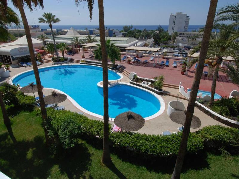 Rundlicher Pool mit bläulichem Wasser, umgeben von Palmen und Liegestühlen am Meer.