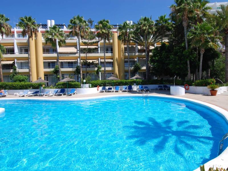 Großer blauer Pool vor einem gelb-beigen Hotel mit Palmen und Sonnenliegen am Beckenrand.