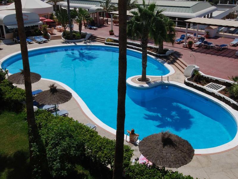 Schwimmbad mit blauem Wasser, umgeben von Palmen, Liegestühlen und rotem Pflaster.