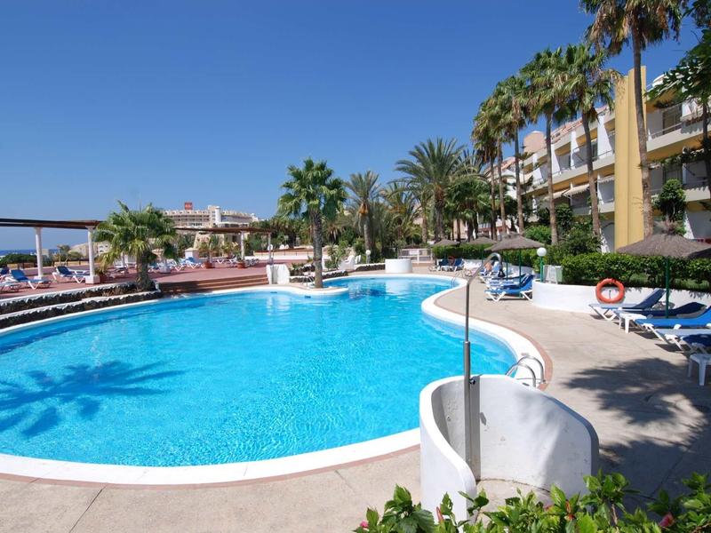 Ein großer, blauer Pool umgeben von Liegestühlen, Palmen und einem Hotelgebäude bei klarem Himmel.