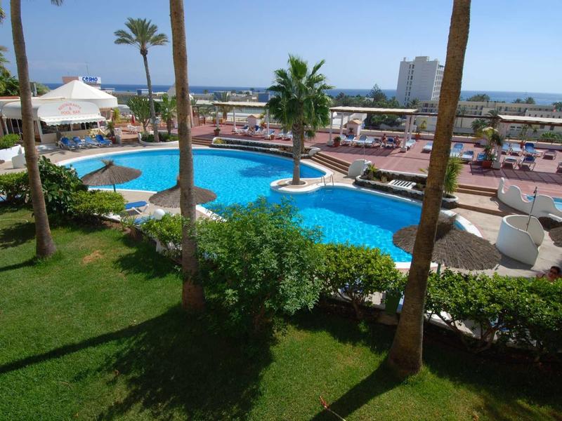 Schwimmbad mit blauer Wasserfläche, umgeben von Palmen, grünen Rasenflächen und Liegestühlen.