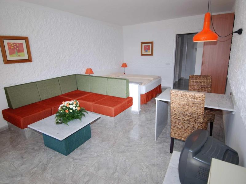 Gemütliches Zimmer mit L-förmiger Couch, Blumen auf Tisch, Bett und orangener Deckenlampe.