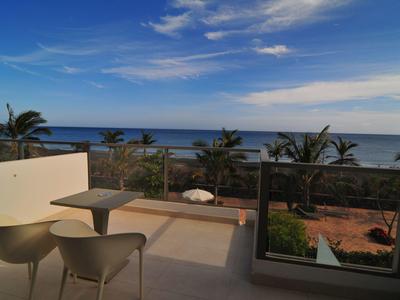 Balkon mit Stühlen, Tisch, Glasgeländer und Blick auf Palmen und Meer unter blauem Himmel.