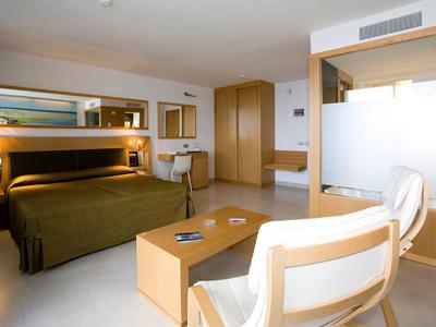 Helles Hotelzimmer mit großem Bett, hellen Möbeln, zwei Sesseln und moderner Beleuchtung.