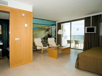 Helles, modernes Hotelzimmer mit Doppelbett, zwei Sesseln, großem Fenster und Balkon mit Meerblick.