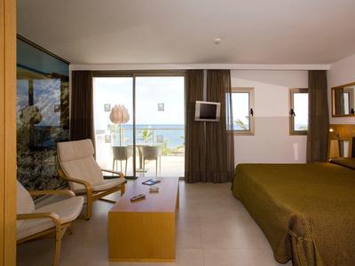Modernes Hotelzimmer mit Doppelbett, zwei Sesseln, Holztisch und Balkon mit Meerblick.