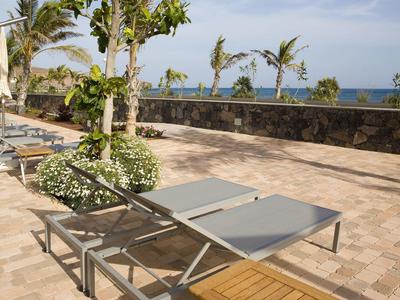 Leerer Liegestuhl vor Palmen, blauer Himmel und Meer im Hintergrund auf gepflasterter Terrasse.
