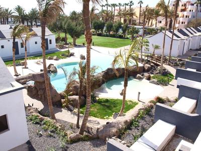 Blick auf weißen Hotelpark mit Pool, Palmen und grünen Rasenflächen an sonnigem Tag.