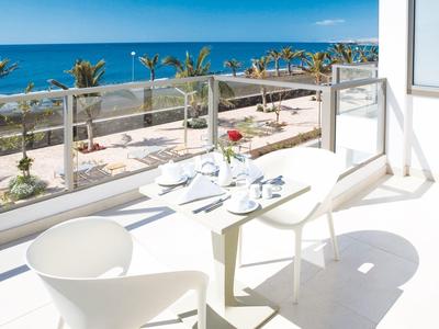 Terrasse mit weißem Tisch und Stühlen, Blick auf das blaue Meer und Palmen unter klarem Himmel.
