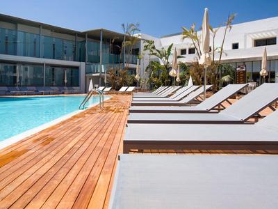 Moderner Poolbereich mit Liegestühlen, Holzdeck und Glasfassade unter blauem Himmel.