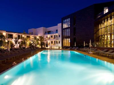 Beleuchteter Pool vor modernem Hotelgebäude bei Nacht mit Liegestühlen am Rand.
