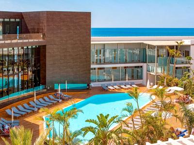 Moderner Hotelpool mit blauer Wasserfläche, Sonnenliegen, Palmen und Glasfenstern bei klarem Himmel.