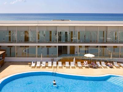 Moderner Hotelpool mit blauem Wasser, umgeben von Liegestühlen und Glasbalkonen eines Gebäudes.