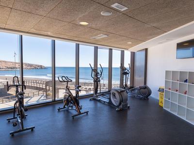 Heller Fitnessraum mit großen Fenstern, Blick aufs Meer, Trainingsräder, Rudergerät und Regalfach.
