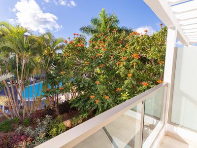 Balkon mit Glasgeländer, Palmen und bunten Blumen bei sonnigem Himmel und blauem Pool.