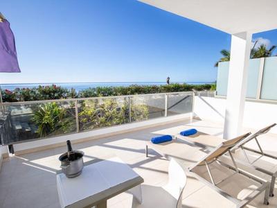 Terrasse mit Liegestühlen, Tisch, Eis und Weinglas, Blick auf Palmen und blaues Meer im Hintergrund.