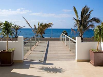 Ein heller Holzsteg führt von einer Terrasse mit zwei Pflanzen im Topf zum blauen Meer mit Palmen im Hintergrund.