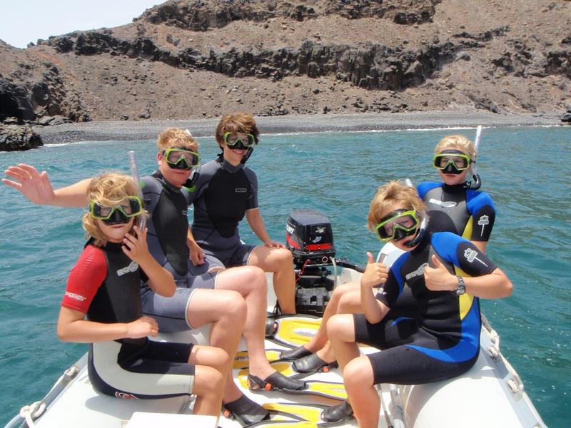 Groep van vijf mensen met snorkeluitrusting op een boot in helder blauw water bij rotsachtige kust.