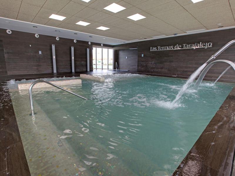 Overdekt zwembad met waterstralen, bruine tegelwanden en grote ramen.