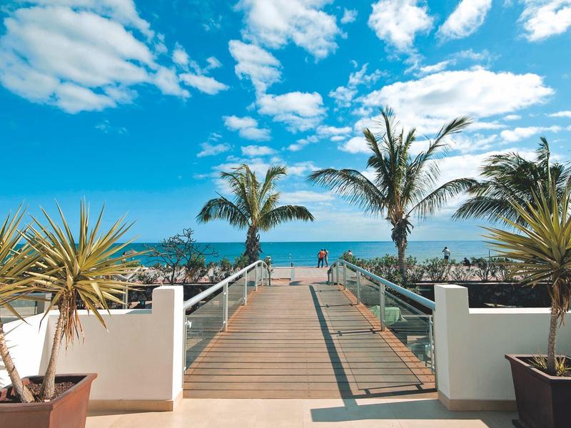 Houten pad naar oceaanzicht met palmbomen onder een heldere blauwe lucht met wolken.