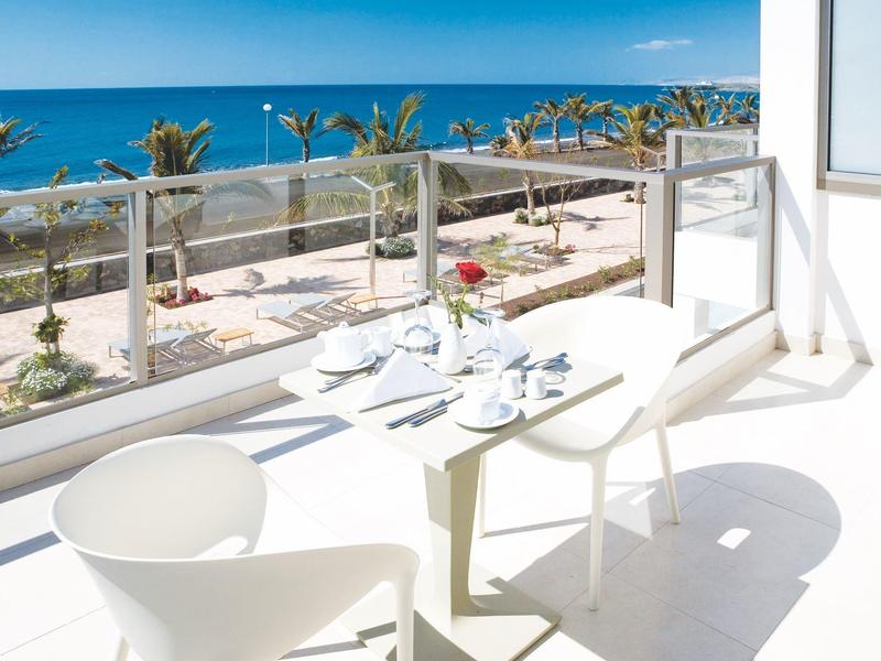 Zonnig balkon met witte tafel en stoelen met uitzicht op de waterkant met palmbomen en blauwe zee.