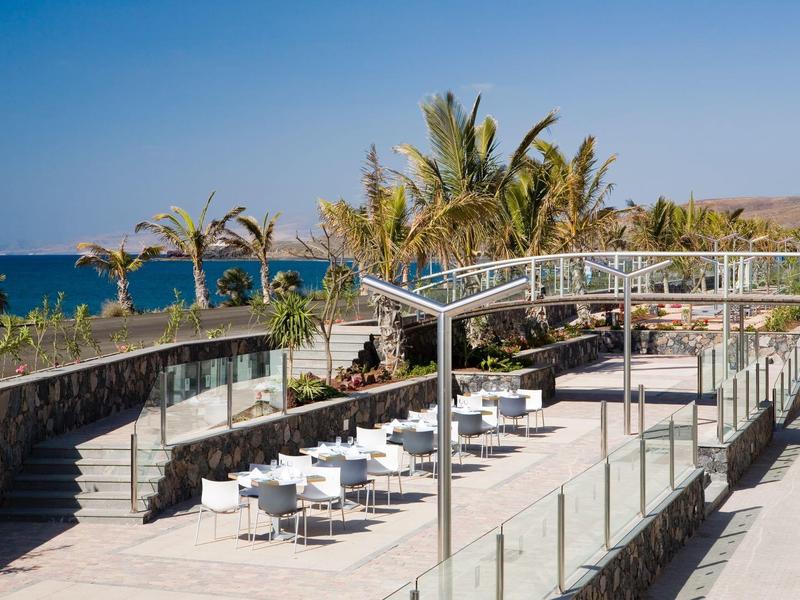 Buiten eetgedeelte met witte tafels en stoelen met uitzicht op de oceaan en palmbomen.