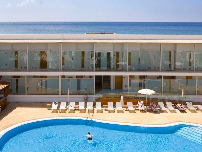 Hotel met een gebogen buitenzwembad, ligstoelen en uitzicht op de oceaan op de achtergrond.