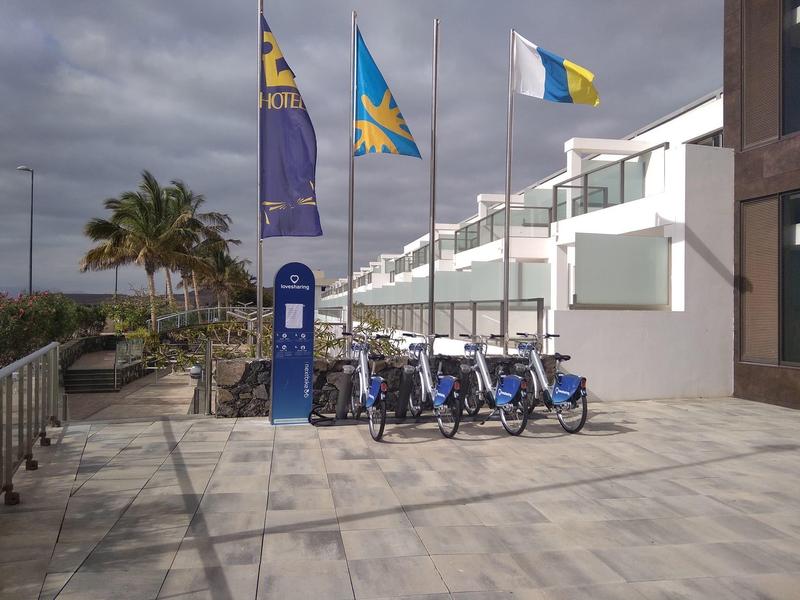Fietsenverhuurlocatie met blauwe fietsen en vlaggen bij een modern wit gebouw en palmbomen.