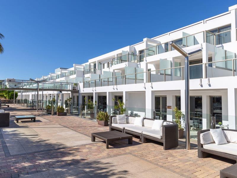 Modern wit hotelgebouw met glazen balkons en buitenzitplaatsen onder een heldere blauwe lucht.