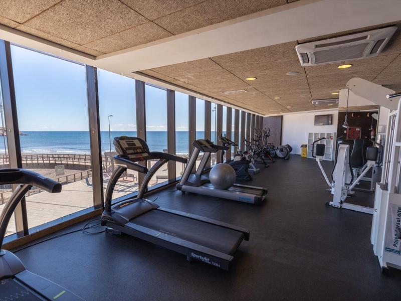 Moderne fitnessruimte met loopbanden en fitnessapparaten bij grote ramen met uitzicht op de oceaan.