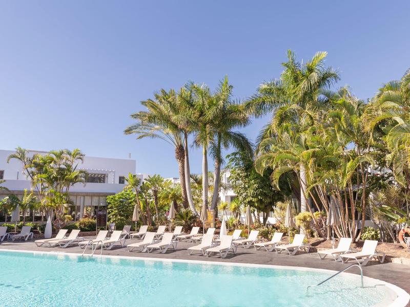 Zwembad met ligbedden en palmbomen onder een heldere blauwe lucht bij een hotel.