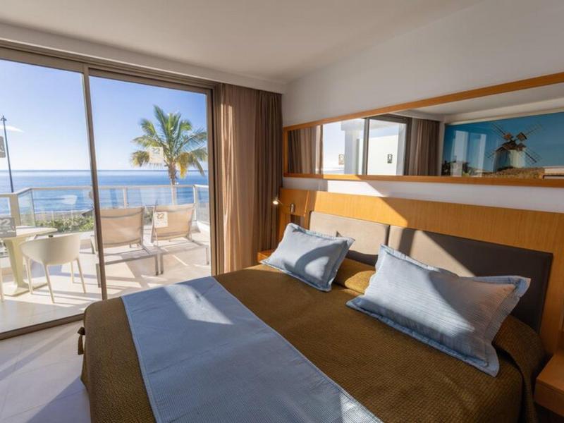 Hotelkamer met tweepersoonsbed, blauwe en bruine lakens, balkon met uitzicht op de oceaan en palmboom.