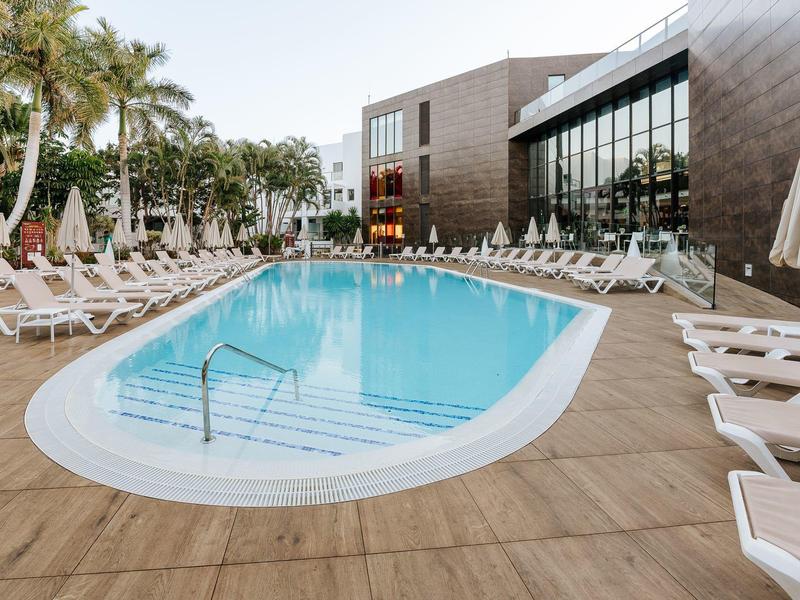 Modern hotelzwembad met witte ligstoelen en palmbomen in een goed onderhouden buitenruimte.
