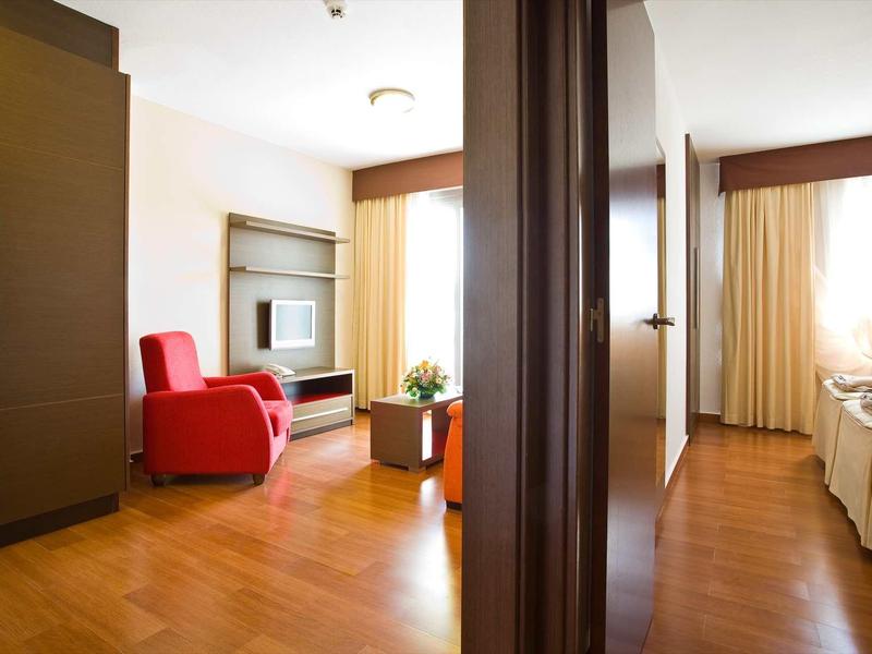 Bright hotel rooms with wooden floor, red armchair, and double bed, separated by a door.