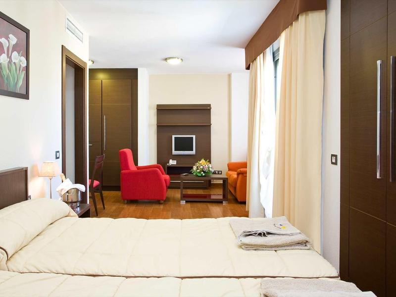 Modern hotel room with large bed, armchairs, and TV in warm colors.