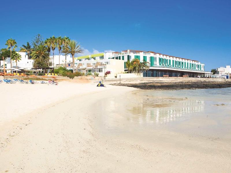 Bright sandy beach with loungers and large hotel buildings in the background under clear blue sky.