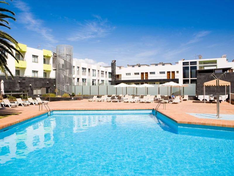 Large outdoor pool in front of a white hotel building under clear blue sky.