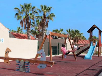 Kinderspielplatz mit Wippe, Schaukel und Rutsche, umgeben von Palmen und blauen Himmel.
