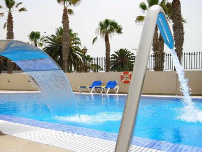 Schwimmbad mit Wasserspielen, blauen Liegestühlen und Palmen im Hintergrund bei Tageslicht.